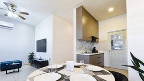 Uma área de refeições com uma mesa redonda posta para duas pessoas, um ventilador de teto e uma cozinha ao fundo.