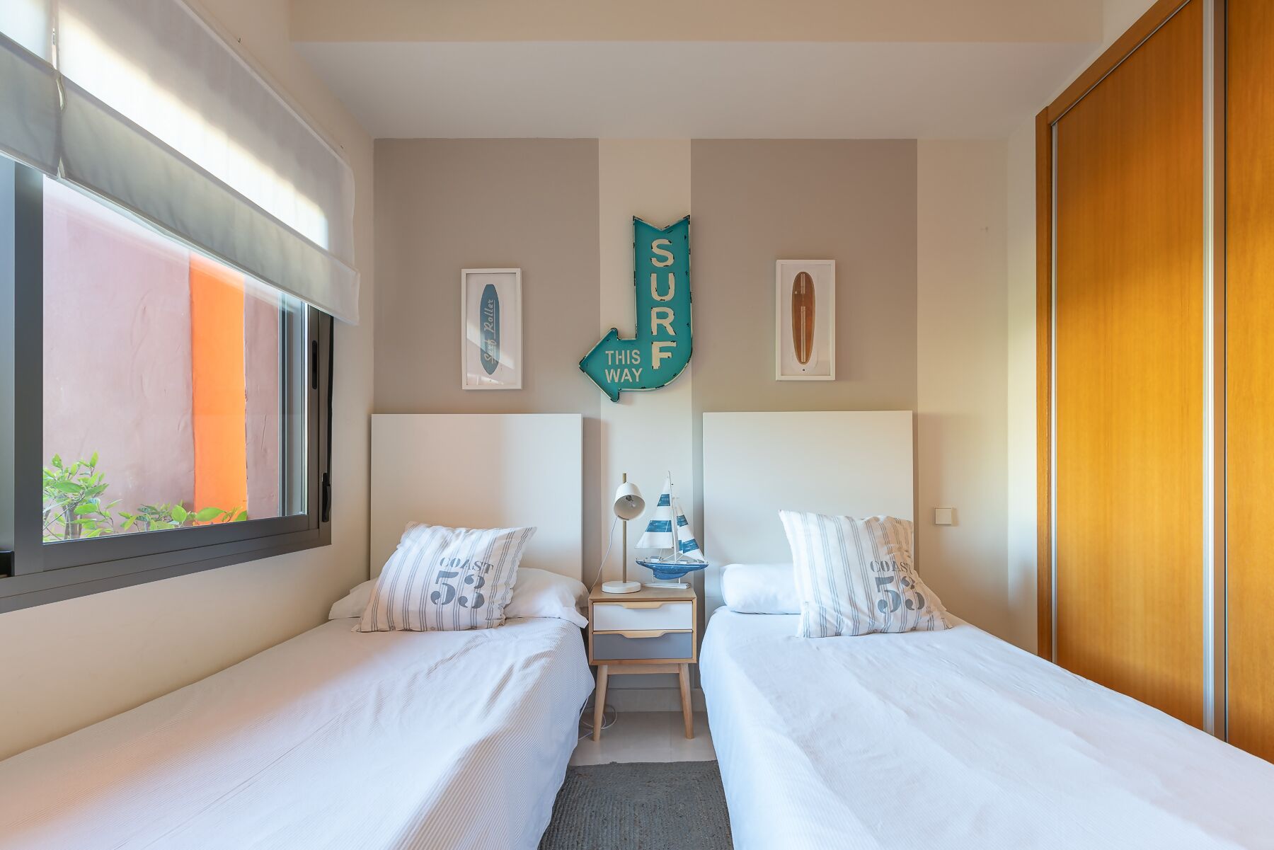 A hotel room with two beds, a window with a view, a surf-themed sign, and a surfboard picture.