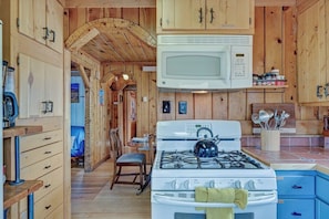 A kitchen with wooden cabinets, a stove, a microwave, and a dining area with a chair and table. - The Cabin Down Below (Buena Vista)