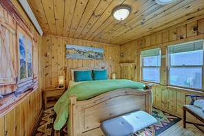 A wooden cabin bedroom with a bed, a bench, a painting, and a window.
