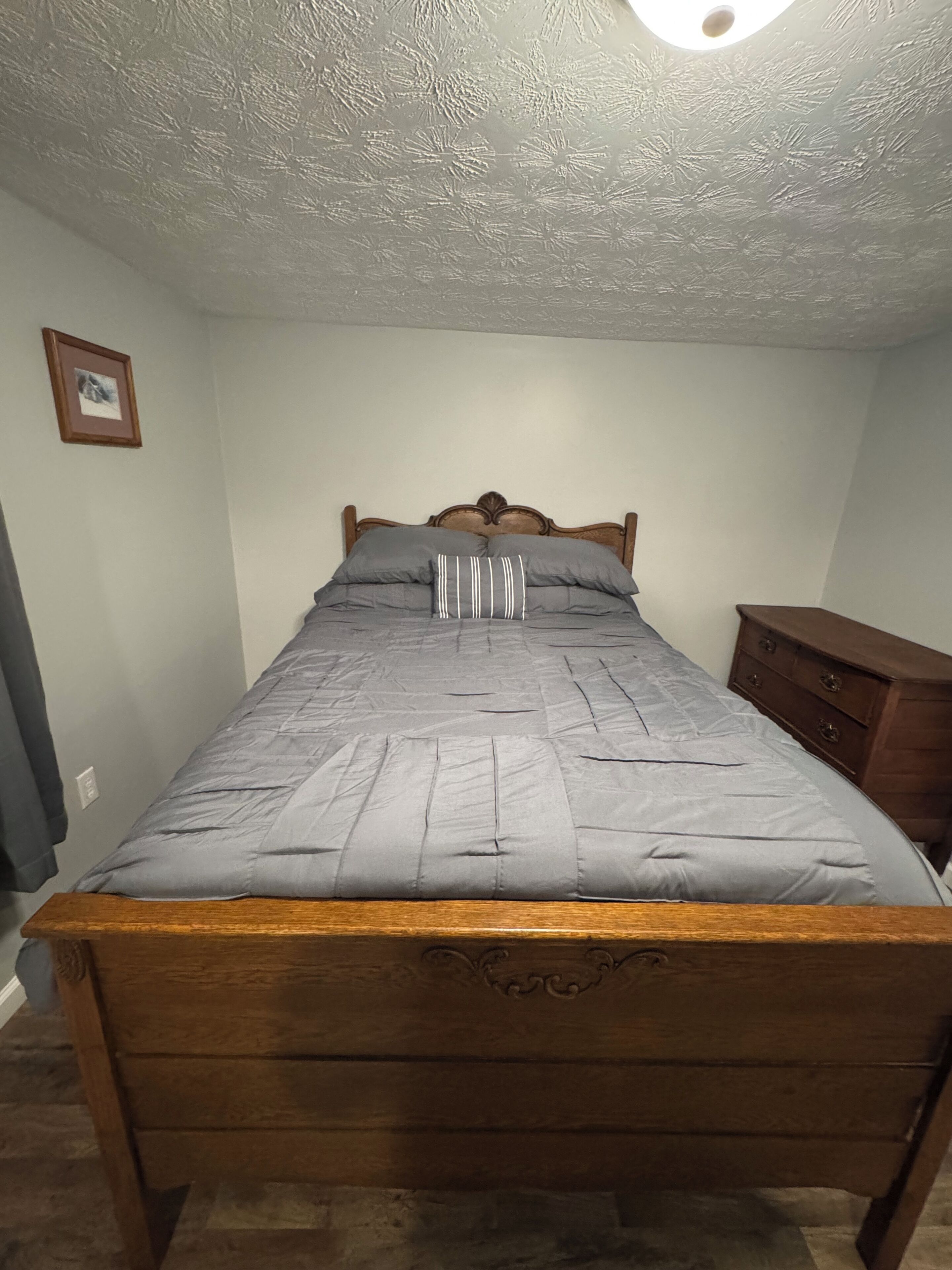 A bedroom with a wooden bed, a nightstand, a framed picture on the wall, and a textured ceiling.