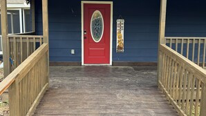 Una terraza de madera que conduce a una puerta roja con un panel de vidrio decorativo, un muro exterior azul y una barandilla de madera.