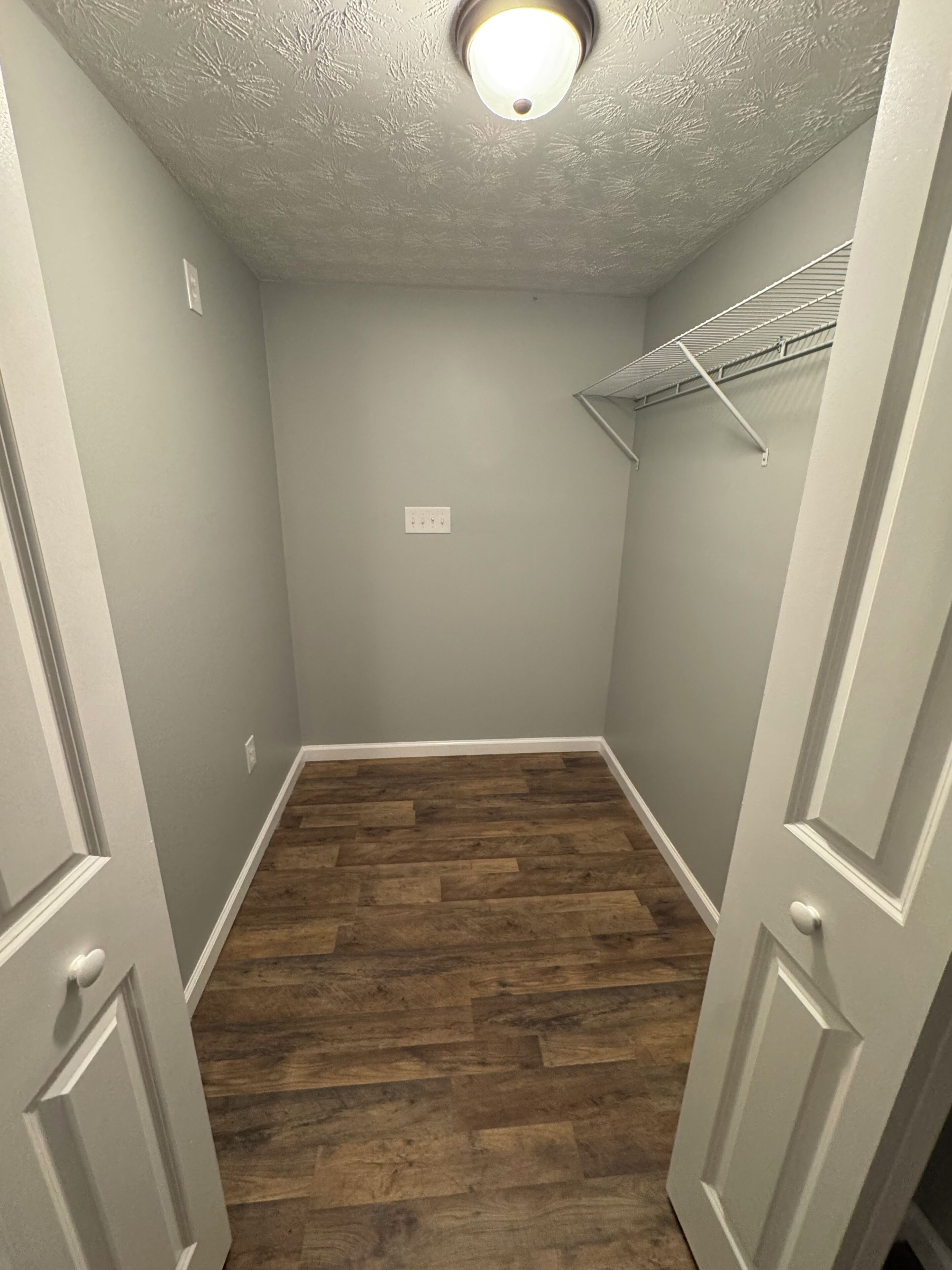 Un couloir étroit avec un plancher en bois, un mur de couleur gris clair et un luminaire au plafond.