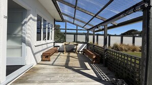 Een overdekt terras met houten banken en een tafel, een wit gebouw met een deur, en een metalen hek.