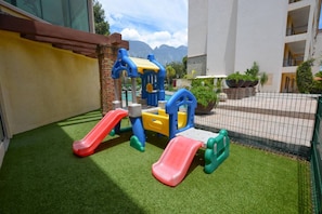 Espaço infantil com escorregadores e estruturas de escalada, cercado por uma grade e vegetação.