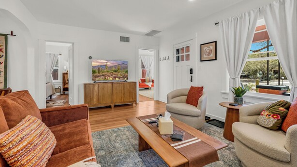 A spacious living room with a sofa, a coffee table, and a TV. There is a door leading to another room and a window with curtains.