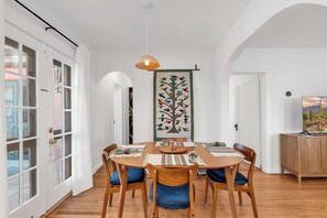 Zona de dinar amb taula de fusta, cadires, lampadari penjant i un quadre penjat a la paret.