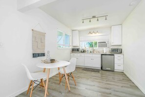 A compact kitchen with white cabinets, a microwave, a toaster, and a dishwasher. There is a round table with chairs and a decorative Buddha statue. - Akaka Falls 6BR Ocean‑View Retreat with Hot Tub (Honomū)