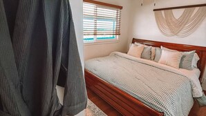 Ein Schlafzimmer mit einem hölzernen Bett, einem Deckenventilator, einem Fenster mit Jalousien und einem Bademantel, der auf einem Gestell hängt.
