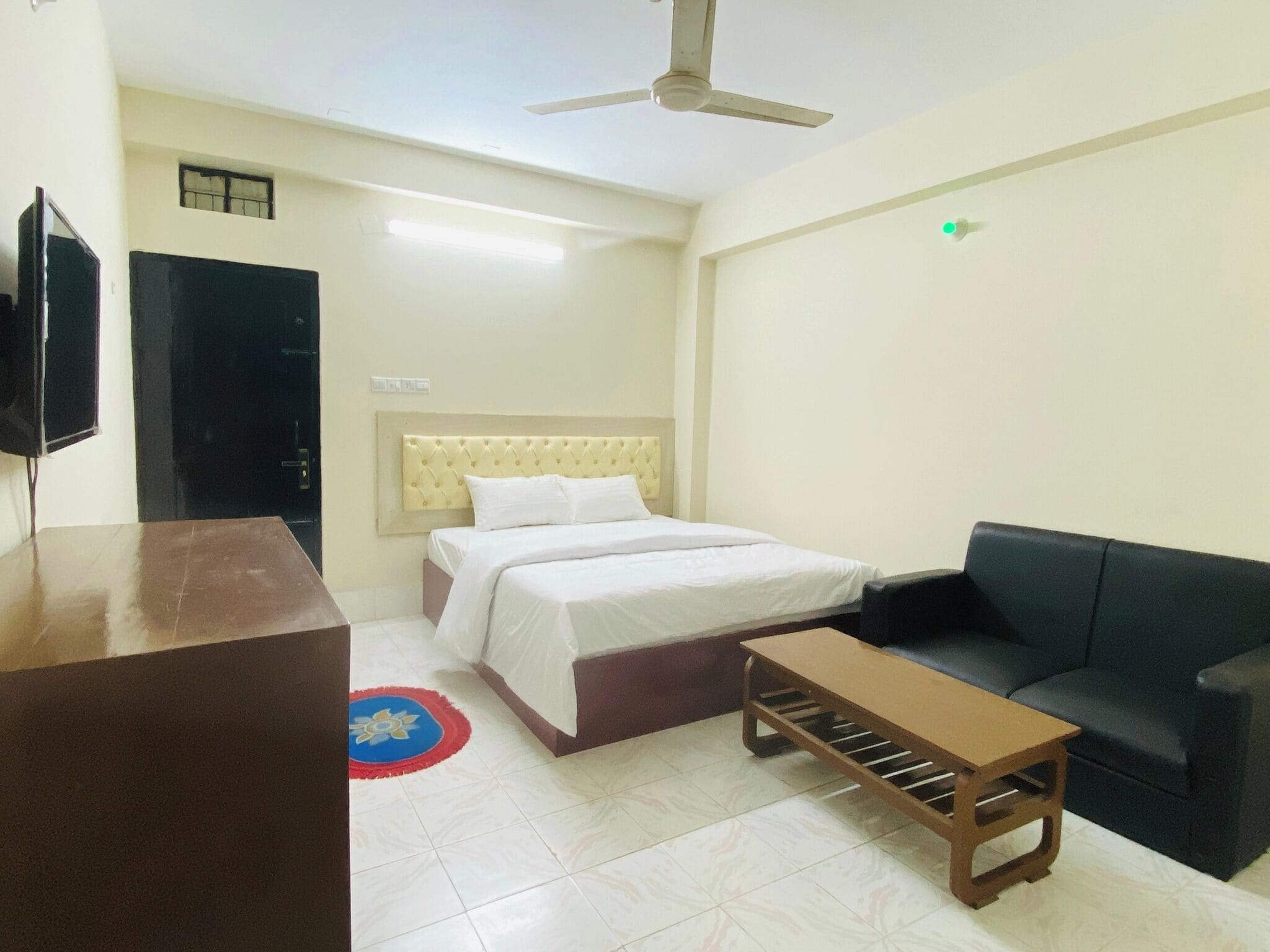 A hotel room with a bed, a sofa, a small table, a ceiling fan, and a television.