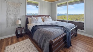Une chambre avec un plafond en bois, un lit avec une tête de lit, une table de chevet, une décoration murale en macramé et de grandes fenêtres offrant une vue dégagée.