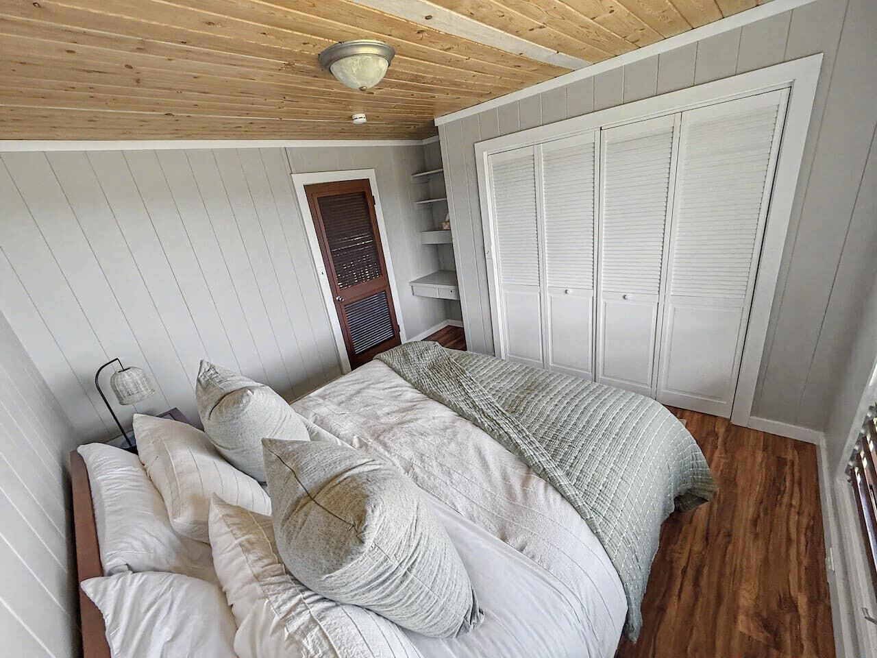 A bedroom with a bed, white walls, wooden flooring, a wardrobe, and a ceiling fan.