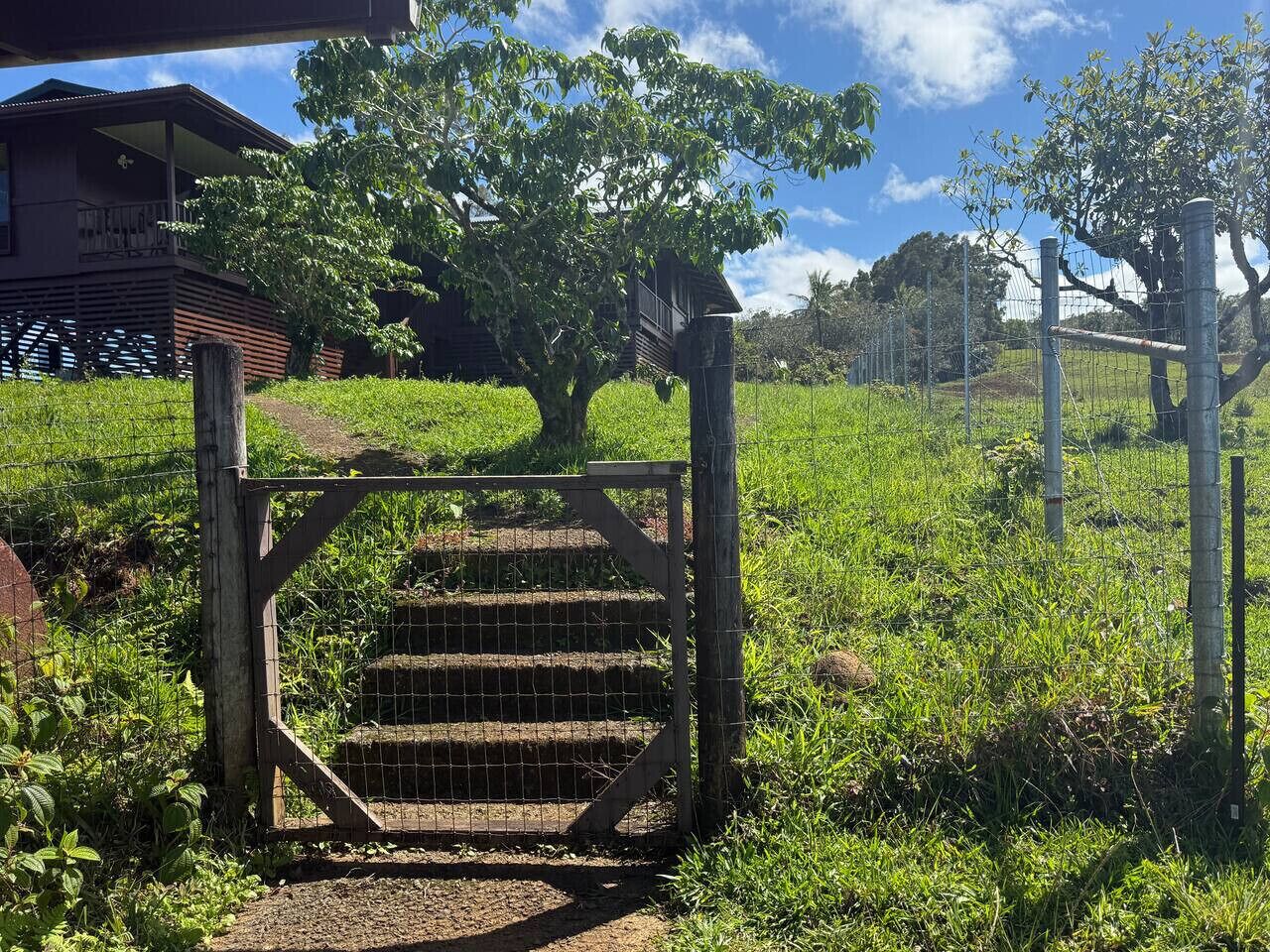 A wooden gate leading to a building with a balcony, surrounded by lush greenery and a clear sky.