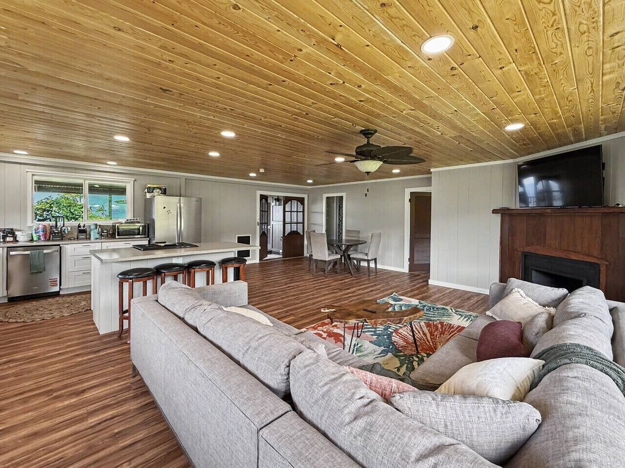 A spacious living area with a wooden ceiling, a kitchen with a white countertop, a dining area with chairs, and a fireplace.