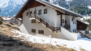 Un chalet a due piani con un balcone in legno, una scala e vista sulle montagne innevate.
