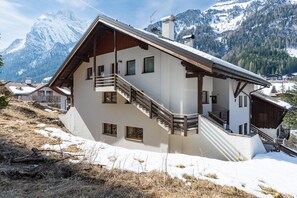 Un chalet de dos pisos con balcón de madera, una escalera y vistas a montañas nevadas.