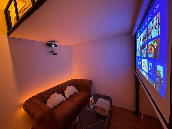 A cozy room with a brown sofa, two plush pillows, a small table with a remote control, a folded towel, and a projector screen displaying various content. - Bugis Social- Newly Renovated (Singapore)
