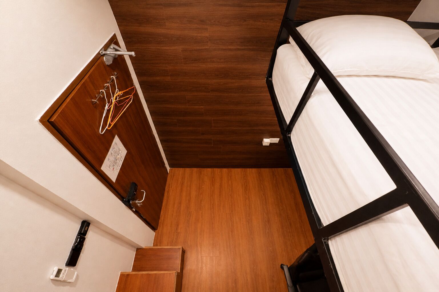 Une chambre avec un lit superposé, un plancher en bois, une porte sur laquelle sont suspendus des vêtements et un petit escalier.