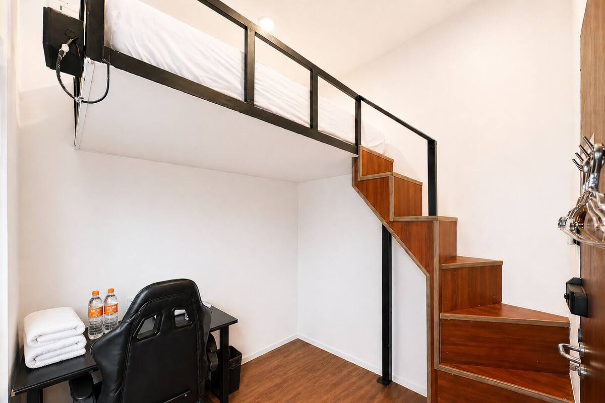 Habitación pequeña con una cama litera, un escritorio con silla, una mesita con botellas y una escalera.