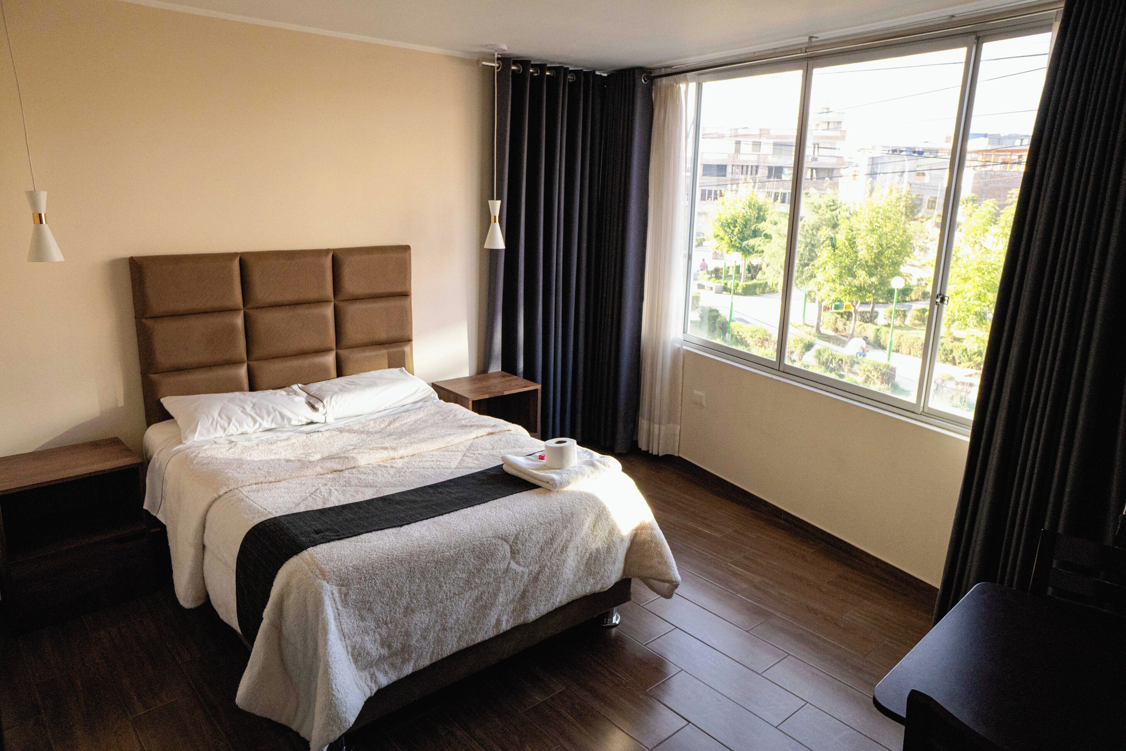 Een hotelkamer met een groot bed, nachtkastje, raam met gordijnen en uitzicht op gebouwen en bomen.