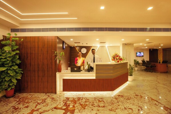 A hotel reception area with a wooden reception desk, a clock, and a TV screen. - Hotel Laals Inn Grand (Ernakulam)