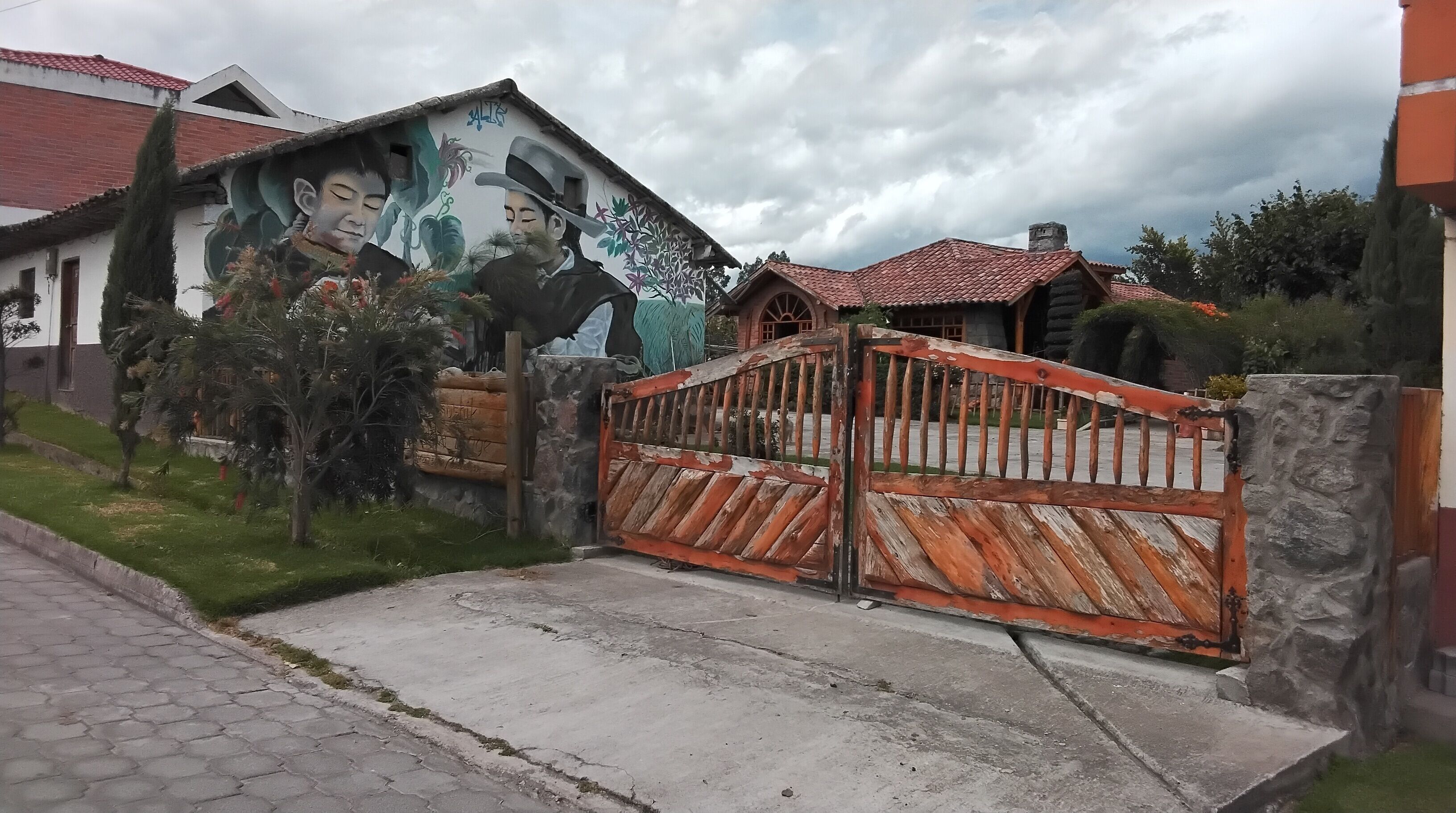 A gated entrance with a mural, a stone wall, and a house with a red-tiled roof.