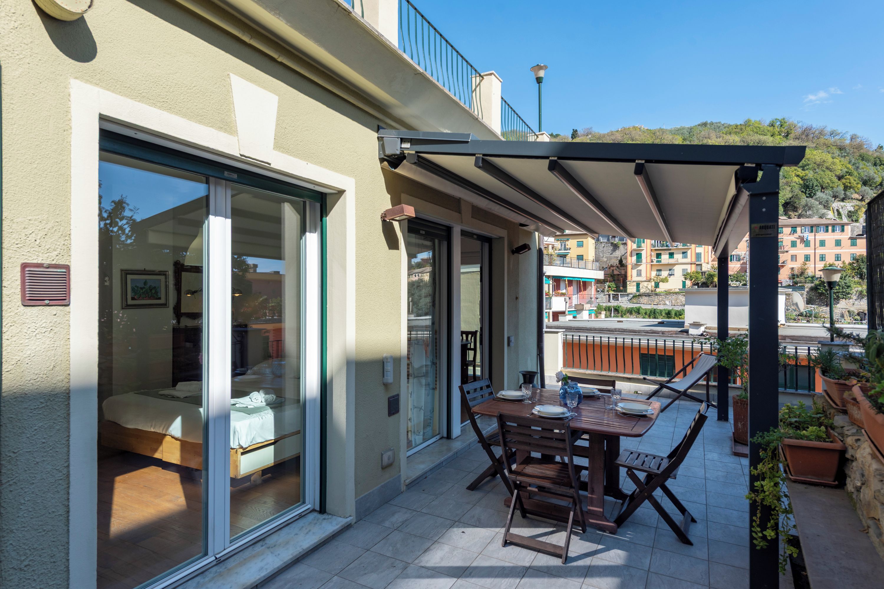 A balcony with a table and chairs, a sliding glass door leading to a bedroom, and a view of buildings in the background.