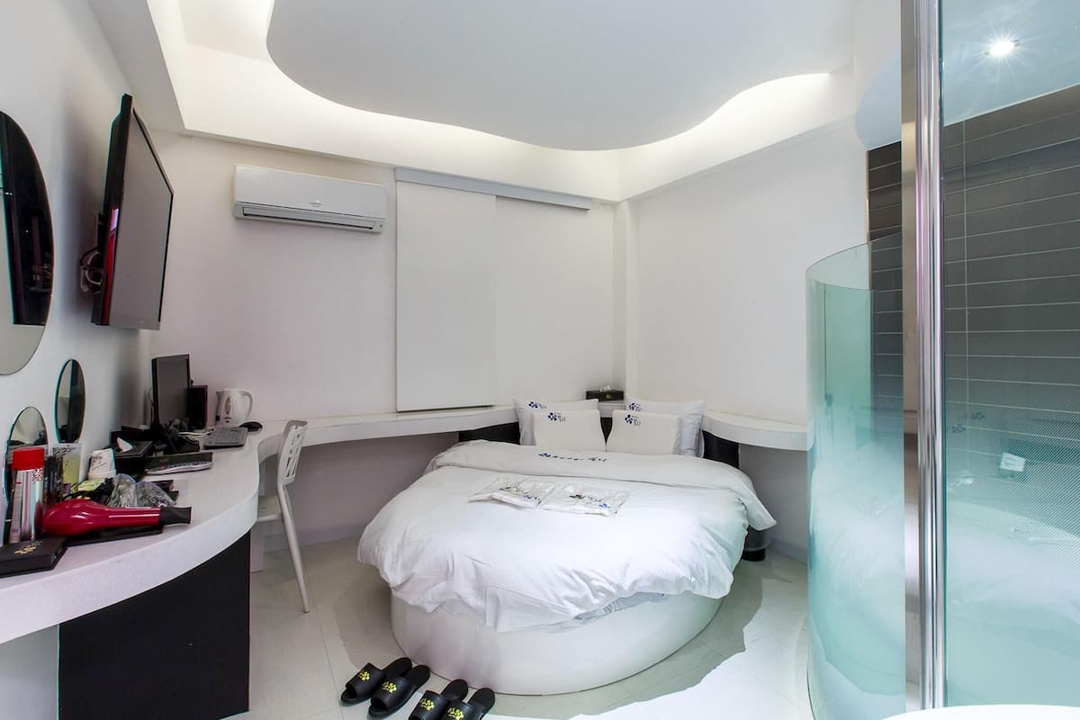 Una habitación de hotel moderna con una cama curvada, cabina de ducha de vidrio, un escritorio blanco con una computadora y un televisor montado.