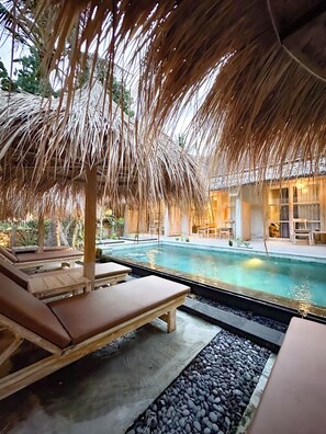 A poolside area with lounge chairs, a thatched roof, and a clear pool.