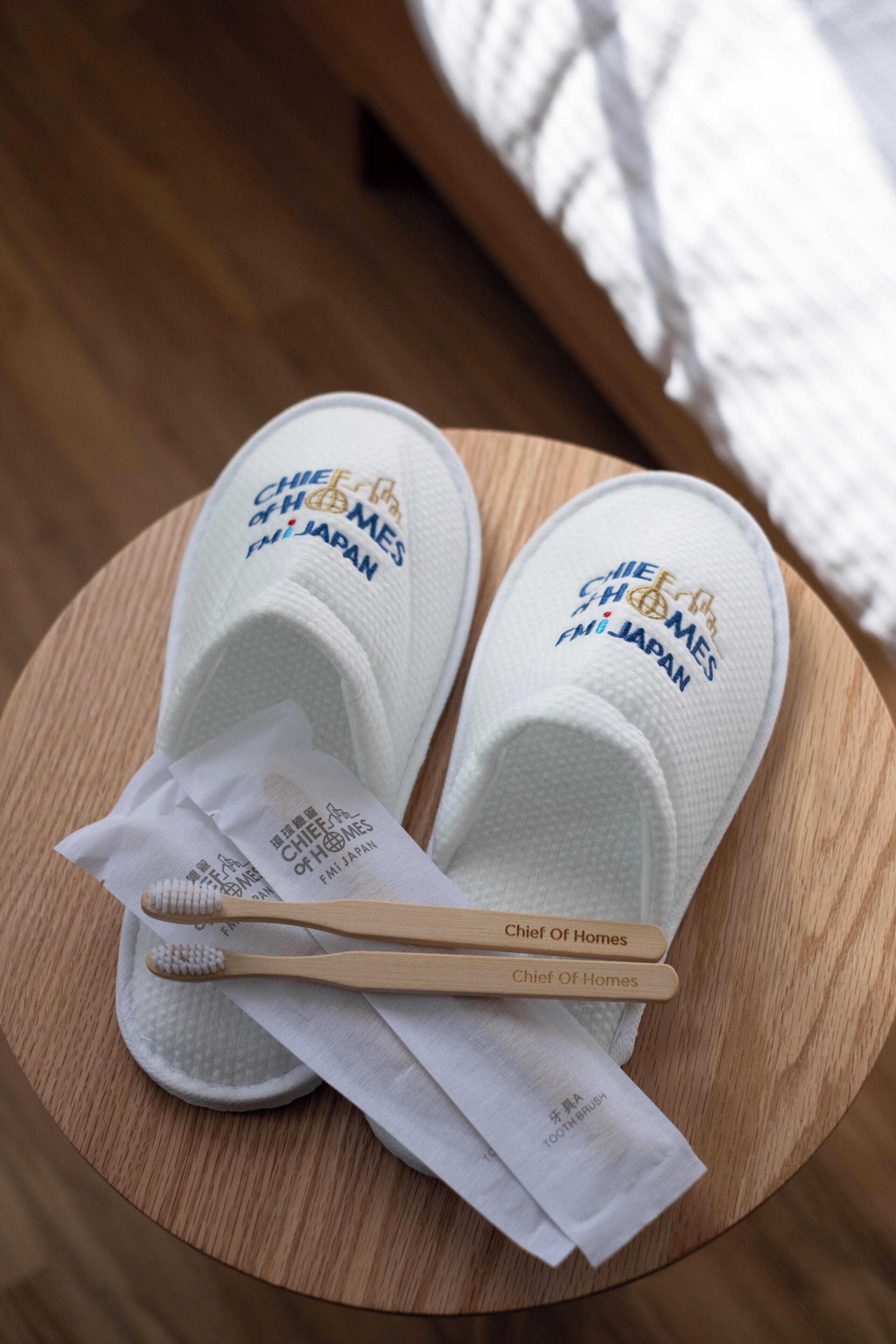 A pair of slippers with 'Chief Of Homes' branding, a pair of wooden toothbrushes, and a white tissue on a round wooden tray.