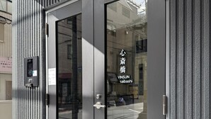 Ein moderner Hauseingang mit Glastür, einem Metall-Sicherungsfach und einem Schild mit japanischen Schriftzeichen.
