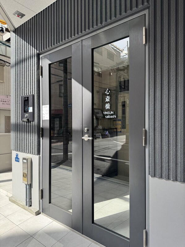 A modern building entrance with a glass door, a metal security box, and a sign with Japanese characters. - WINSUN Shinsaibashi (Osaka)