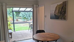 Una habitación con una mesa redonda de madera, dos sillas negras, un ventanal con cortinas, un cuadro en la pared y vistas a un jardín y zona de estar exterior.