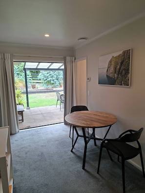 Una habitación con una mesa redonda de madera, dos sillas negras, un ventanal con cortinas, un cuadro en la pared y vistas a un jardín y zona de estar exterior.