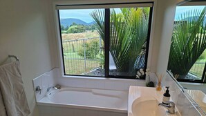 Un baño con bañera, lavamanos, espejo y ventana con vistas a zonas verdes y colinas.