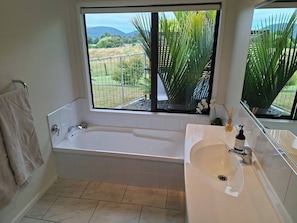 Un baño con bañera, lavamanos, espejo y ventana con vistas a zonas verdes y colinas.