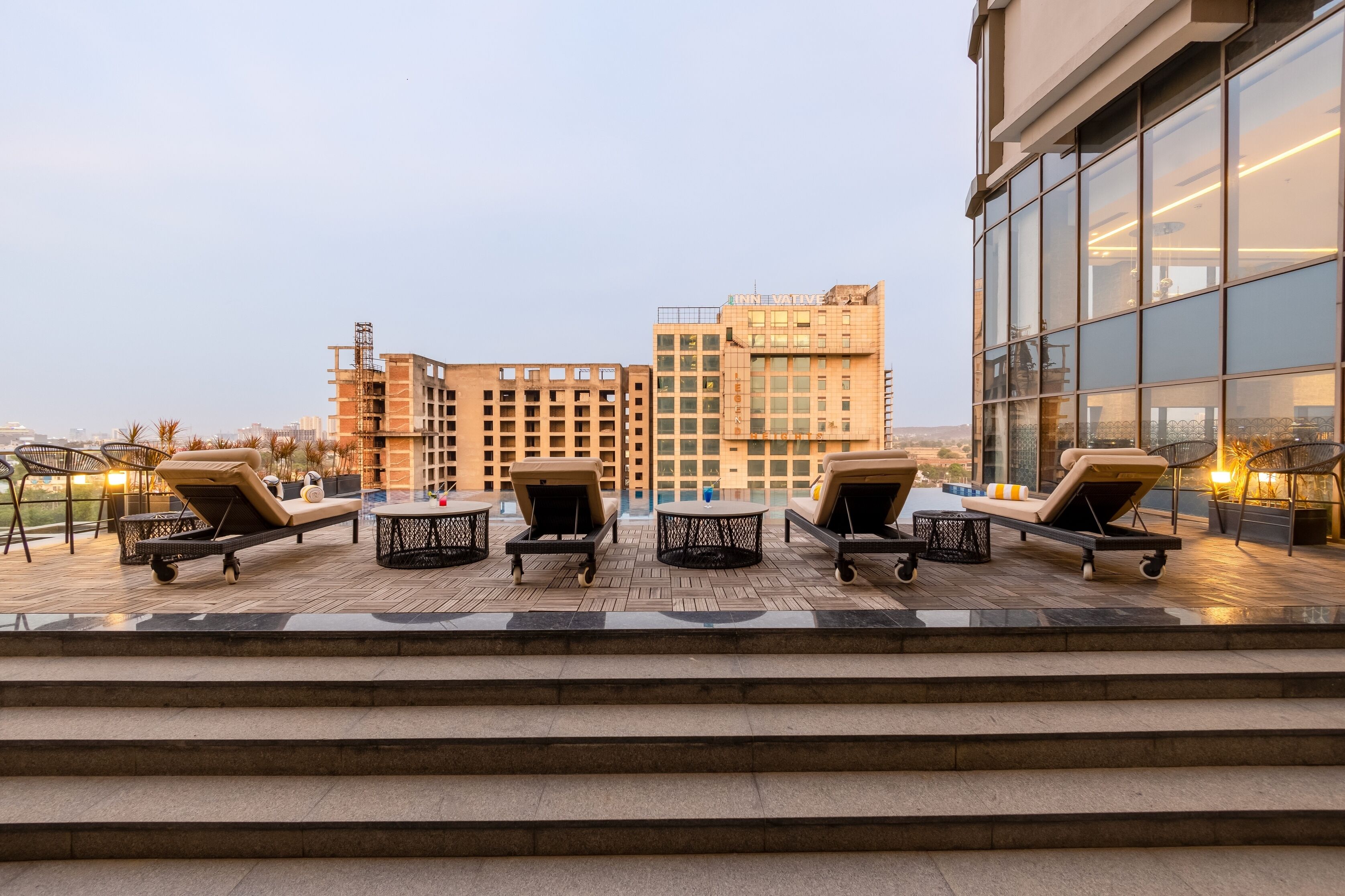 A rooftop lounge area with sun loungers and a pool, overlooking a cityscape with modern buildings.