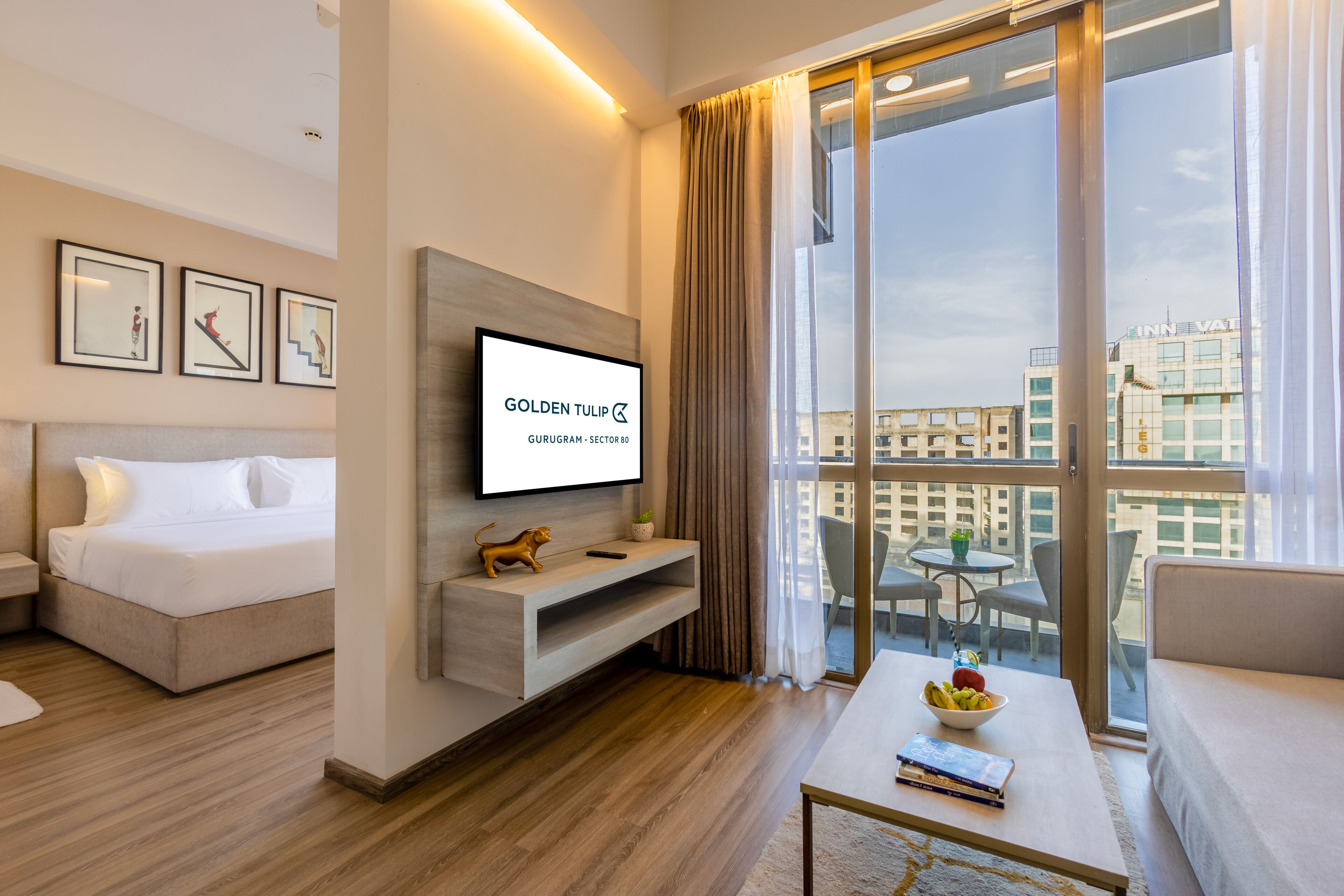 A modern hotel room with a bed, a TV mounted on the wall, a balcony with a view of the city, and a dining area with a table and chairs.