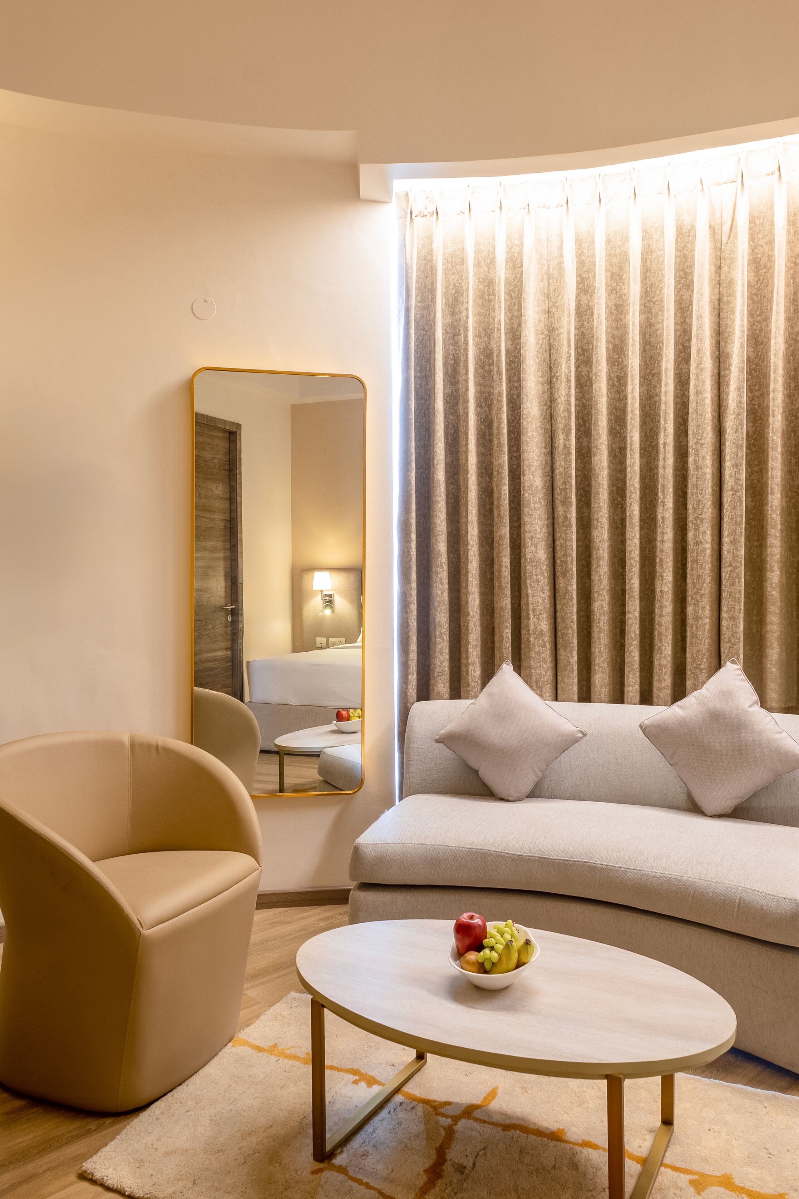 A modern living room with a sofa, armchair, round coffee table with fruit, and a large mirror.