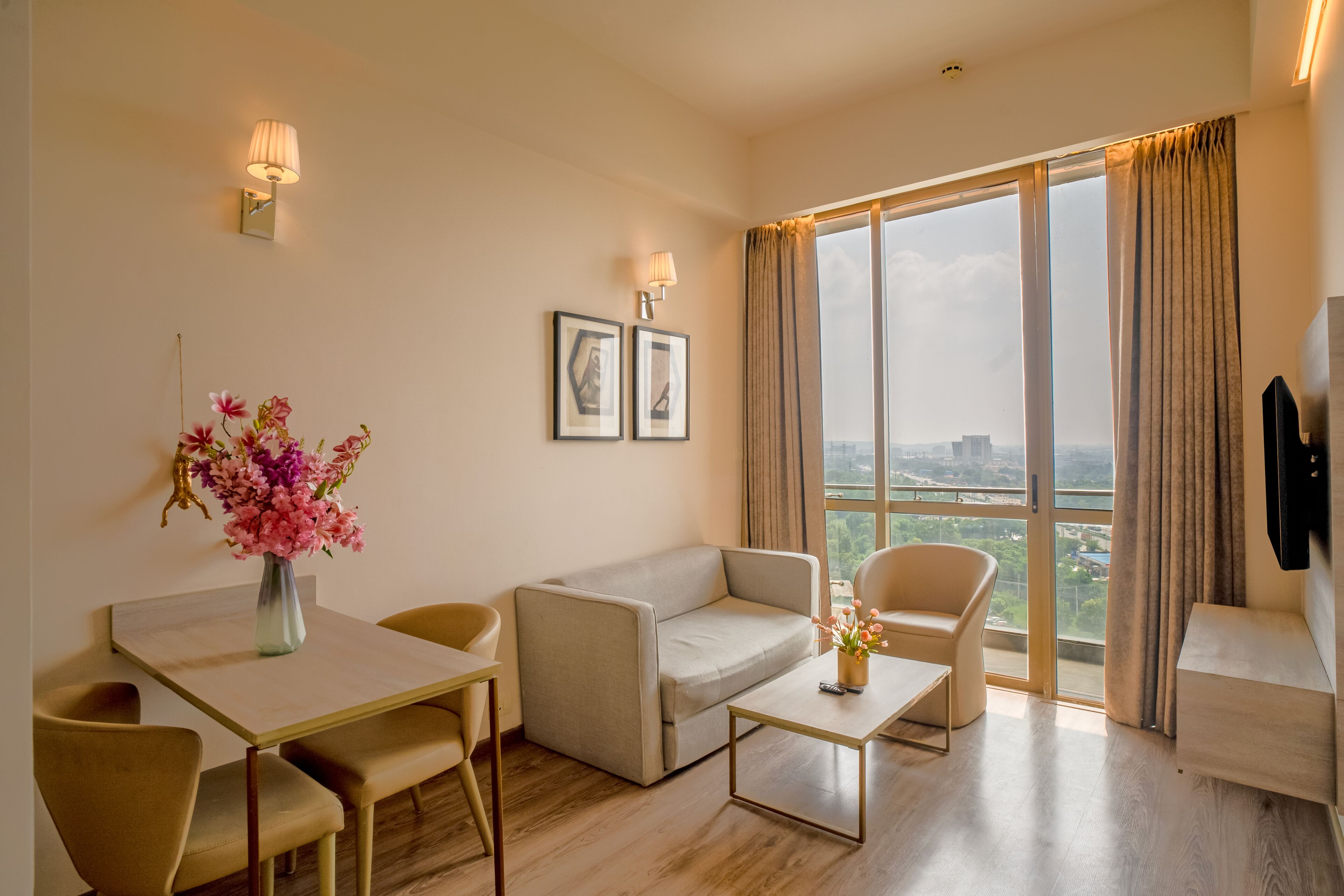 Una sala de estar moderna con sofá, sillones, una mesa de centro y una mesa de comedor. Hay dos lámparas de pared, un jarrón con flores y cuadros enmarcados. Un ventanal con cortinas ofrece vistas de la ciudad.