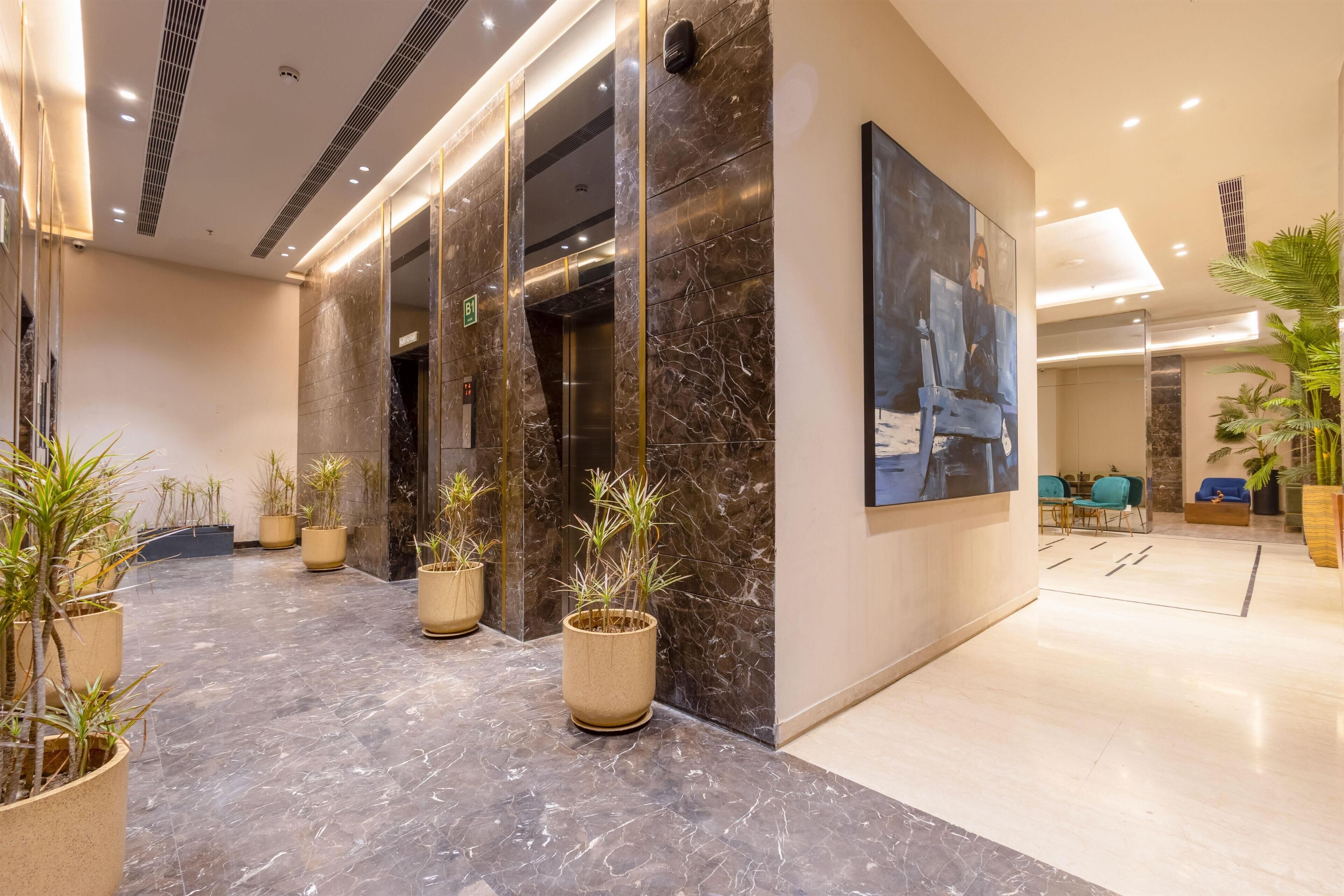 A modern lobby with marble flooring, potted plants, a large abstract painting, and a seating area with blue chairs.