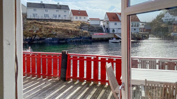 A wooden deck with a red railing, overlooking a calm water body with houses in the background.