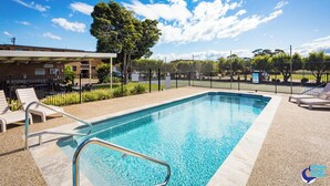 A swimming pool with lounge chairs, a fenced area, and a building in the background. - Apollo Holiday Units (Narooma)