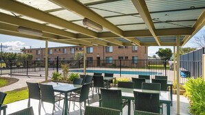 A covered outdoor area with tables and chairs, a pool, and a brick building in the background. - Apollo Holiday Units (Narooma)