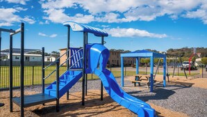 A playground with a blue slide and climbing structure, surrounded by a fence and a picnic table. - Apollo Holiday Units (Narooma)