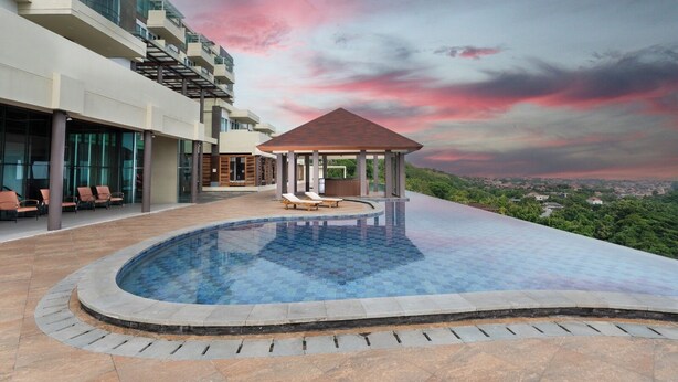 A rooftop infinity pool with lounge chairs and a gazebo, overlooking a cityscape. - Kamala Bali Resort by AGATA (Nusa Dua)