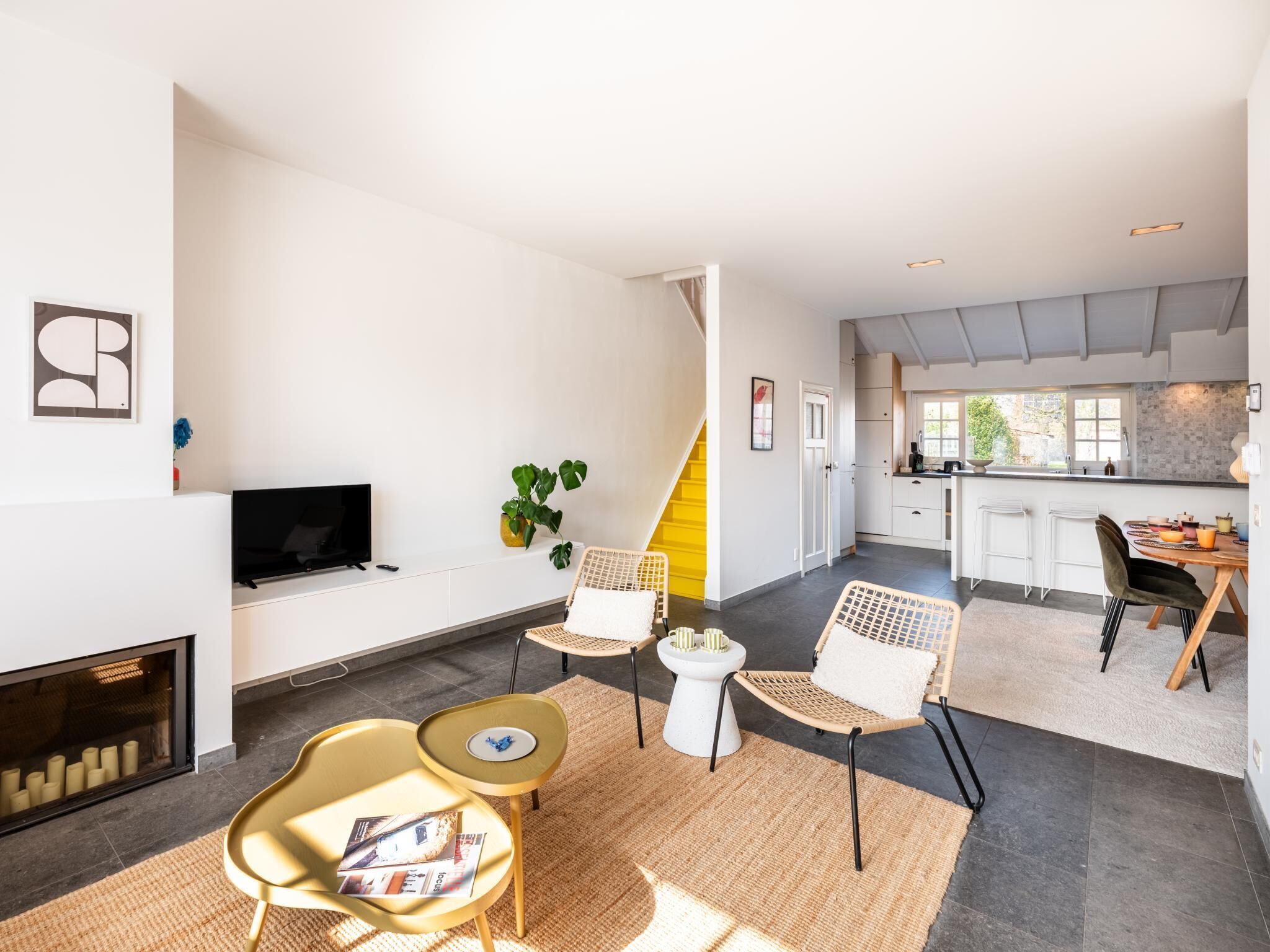 A modern living room with a fireplace, a TV, and a dining area with a kitchen in the background.