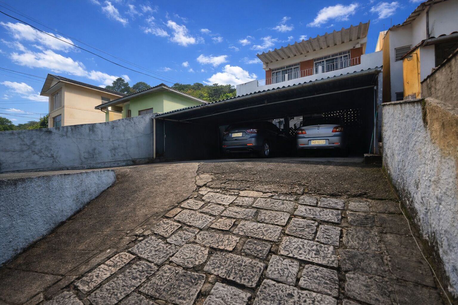 Un garaje residencial con dos coches aparcados en su interior, un camino de entrada empedrado de piedra y una casa de dos pisos con techo plano y un balcón.