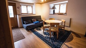 A cozy room with a sofa, a round table, and two chairs. There are two windows with curtains, a lamp on a shelf, and a wooden ceiling.