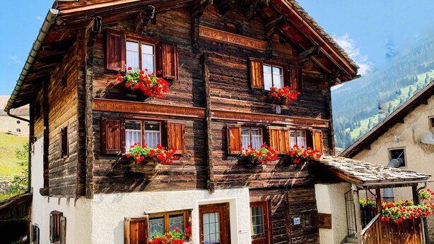 A traditional wooden chalet with multiple windows, balconies with red flowers, and a stone staircase leading to the entrance.
